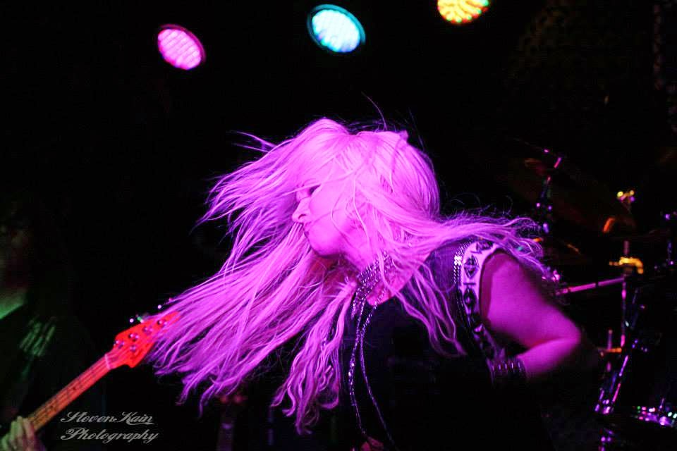 Hunter Storm, The Metal Valkyrie, onstage, soulfully lost in emotion, eyes closed, hair whipping in pink-lit reverie, adorned in studded wristband, black leather motorcycle glove, and spandex muscle shirt with chains and studs. Bass guitar headstock, drum kit cymbals, and vibrant lights in the background.
