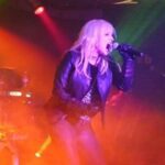 Hunter Storm, The Metal Valkyrie, rocking the stage, screaming intense Metal lyrics, platinum blonde hair flowing in the red-hot lights, adorned in black leather gloves and jacket, zebra shirt, and metallic gray denim.