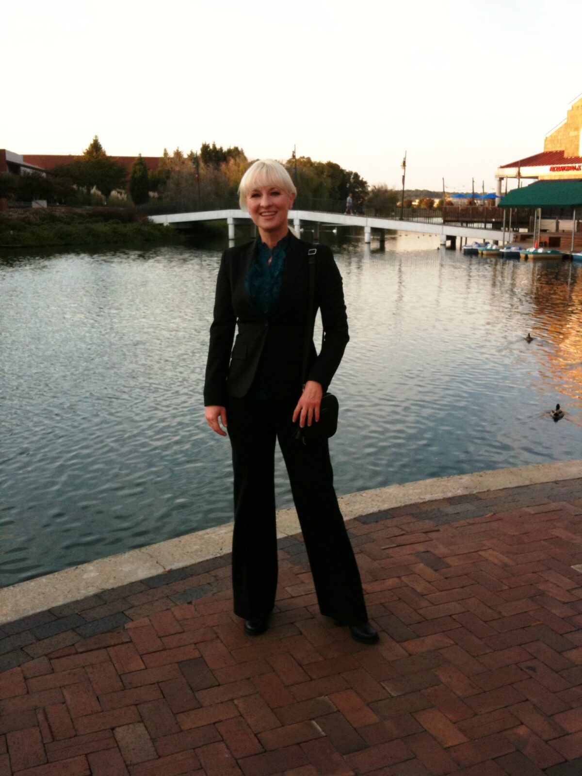 Hunter Storm by a tranquil lake at sunset, epitomizing tech consulting and speaking engagements in a chic, black Theory suit with teal silk designer blouse and Prada boots.
