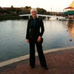 Hunter Storm by a tranquil lake at sunset, epitomizing tech consulting and speaking engagements in a chic, black Theory suit with teal silk designer blouse and Prada boots.