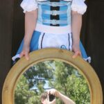 Hunter Storm dressed as Alice in Wonderland, wearing a light blue and white dress, white platform heels, and holding a mirror that reflects the photographer, Ken Diliberto, taking the picture.