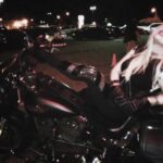Hunter Storm, The Metal Valkyrie, fierce in black spandex and studded leather, reclines on a motorcycle, capturing the rebel spirit under city lights.