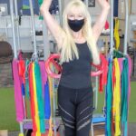 Hunter Storm, The Metal Valkyrie, engaging in therapeutic exercises with Therabands on a physical therapy rack, wearing a black Alo Yoga workout outfit.
