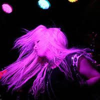 Hunter Storm, The Metal Valkyrie, onstage, soulfully lost in emotion, eyes closed, hair whipping in pink-lit reverie, adorned in studded wristband, black leather motorcycle glove, and spandex muscle shirt with chains and studs. Bass guitar headstock, drum kit cymbals, and vibrant lights in the background.